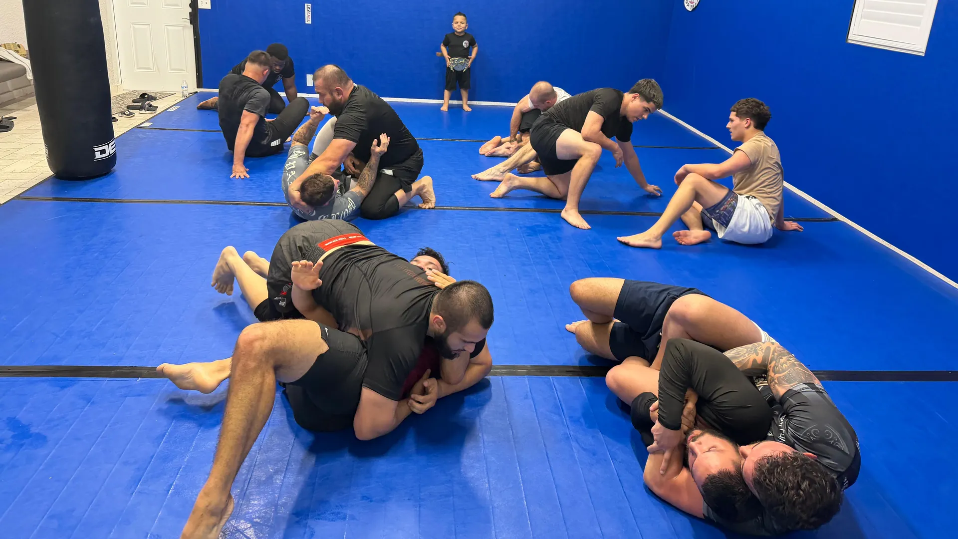 Students rolling and grappling in pairs during an open mat session on blue mats, with a heavy bag and an American Top Team logo on the wall