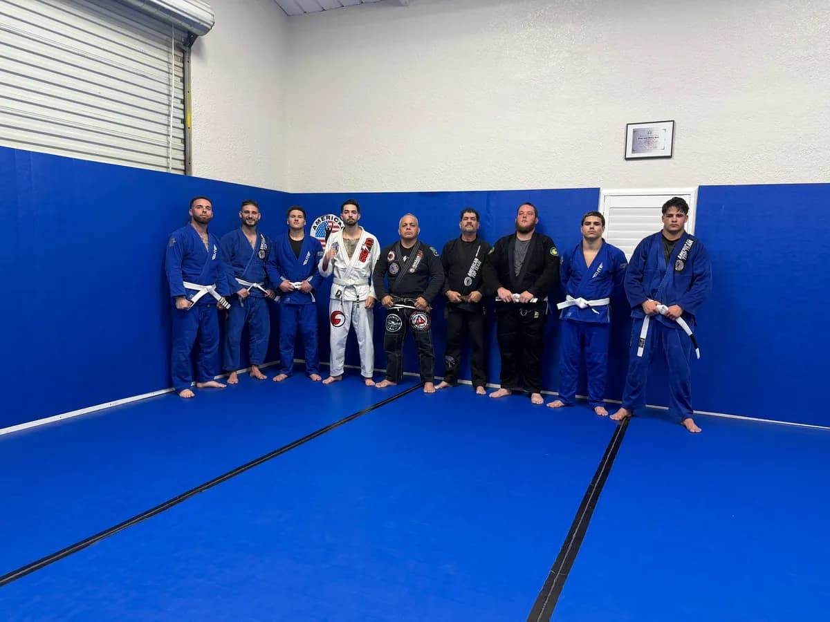 Adult students in blue, white and black gis lined up on blue mats in the corner of the academy, with an American Top Team wall logo