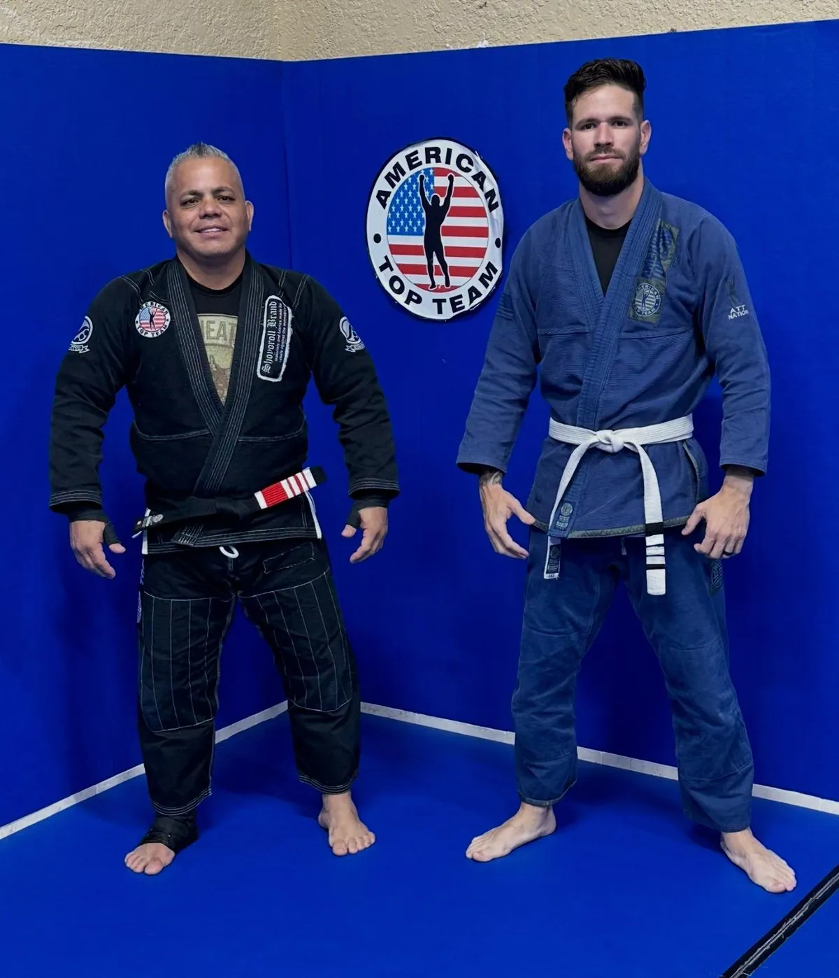 Master Willy Hardoy with white hair in a black gi standing next to a student in a blue gi with a white belt, beside an American Top Team wall logo